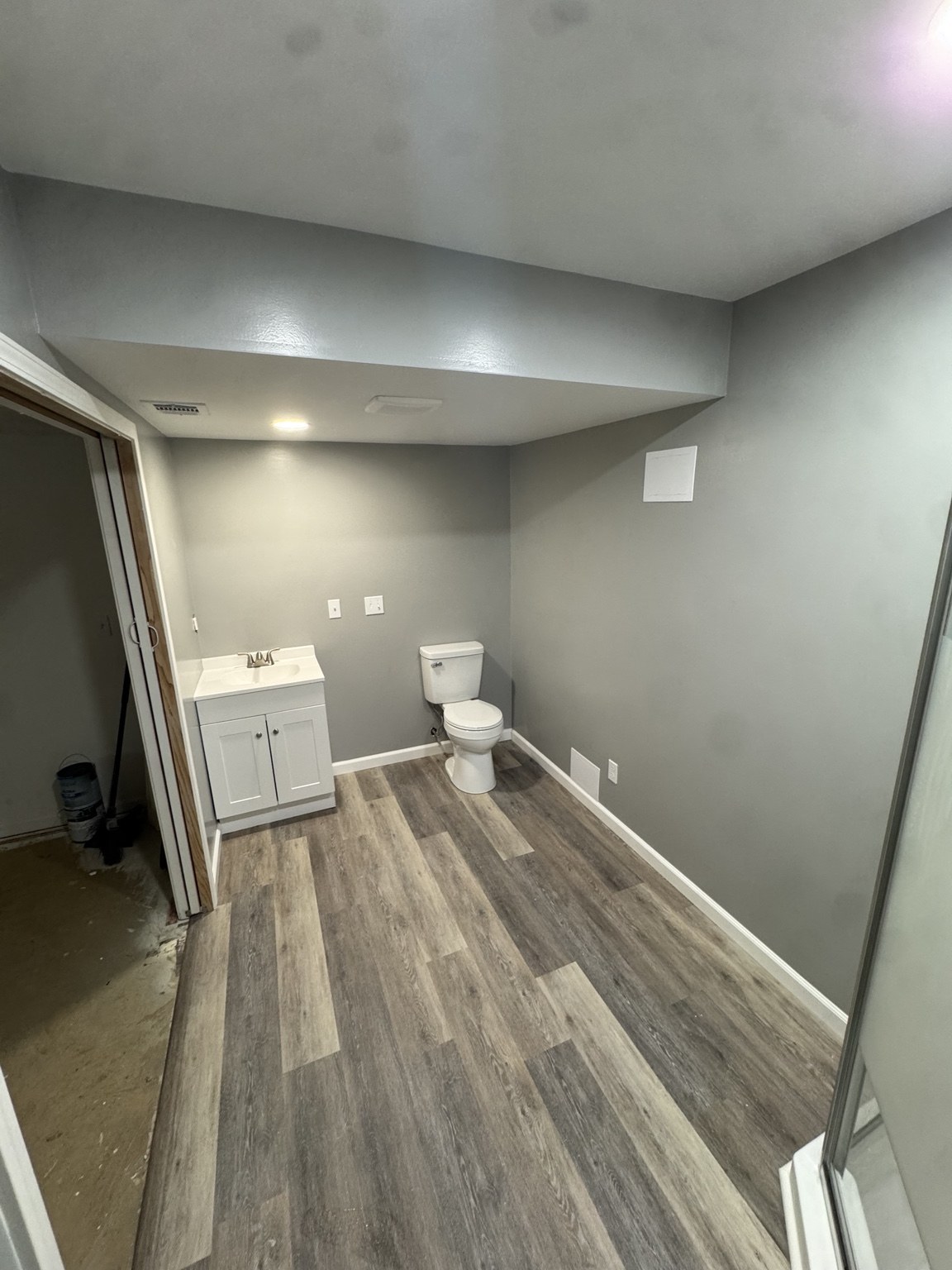 Same bathroom after renovation, with gray walls, white vanity, updated toilet, and wood-look flooring